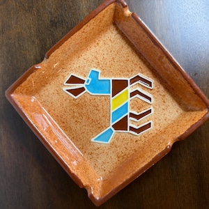 May include: A square ceramic ashtray with a speckled orange and brown glaze. The center features a stylized graphic of a four-legged animal in blue, brown, and yellow, outlined in white, with notches on the corners for resting items.