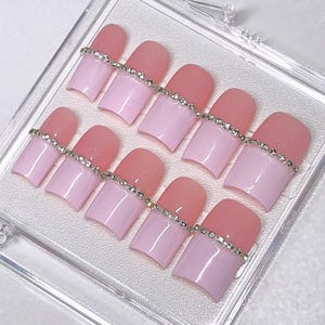 May include: A set of press-on nails in a clear plastic case. Each nail is a square shape with a light pink base and a darker pink tip, adorned with a line of small, sparkling rhinestones. The text "girlypopcouture!" is visible.