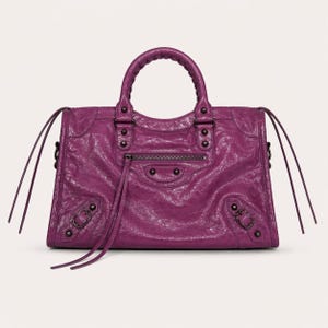 May include: A textured, deep purple handbag with a top handle and a zip closure. The bag features decorative studs, buckles, and long, thin leather straps. The bag has a structured shape and a slightly distressed appearance.