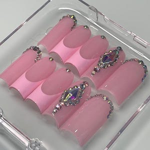 Könnte beinhalten: Ein Set aus rosa Press-on-Nägeln mit French-Tip-Design. Einige Nägel sind mit schillernden Strasssteinen und einem großen Edelstein verziert. Die Nägel werden in einem transparenten Kunststoffbehälter präsentiert. Der Text "girlypopcouture!" befindet sich oben rechts.