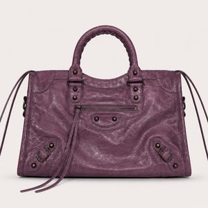 May include: A textured, purple leather handbag with a top handle and a zip closure. The bag features decorative studs, buckles, and long, thin straps. The bag has a classic, structured design.