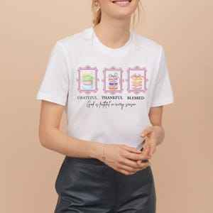 Puede incluir: Camiseta blanca con un diseño que presenta tres ilustraciones enmarcadas de macarons coloridos. Las palabras "Grateful, Thankful, Blessed" están encima de las ilustraciones, y "God is faithful in every season" debajo.