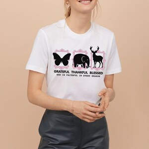 Puede incluir: Camiseta blanca con un diseño de silueta de mariposa, elefante y ciervo dentro de marcos rosas. El texto "GRATEFUL THANKFUL BLESSED GOD IS FAITHFUL IN EVERY SEASON" está impreso debajo. La camiseta tiene mangas cortas.