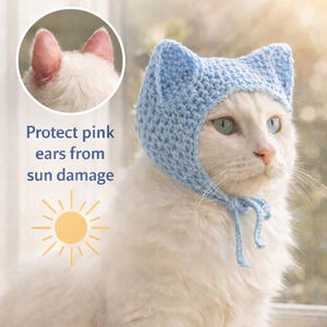 May include: A white cat wearing a light blue crocheted hat with ear-shaped details and a tie. The image includes text that reads "Window Sunburn Protection Cape (For White Cats)" and "Protect pink ears from sun damage". A sun graphic is also present.