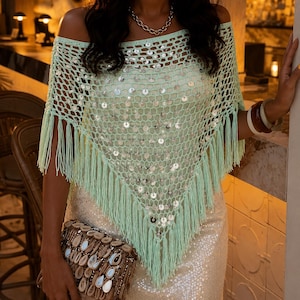 Può includere: Un poncho all'uncinetto color verde chiaro con spalle scoperte, caratterizzato da un motivo a maglia larga, paillettes circolari argentate su tutto il tessuto e lunghe frange lungo i bordi inferiori. Il capo ha una forma triangolare e un ampio scollo.
