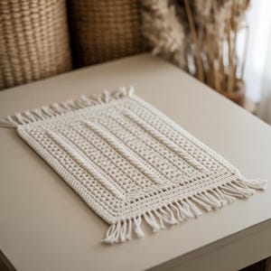 Könnte beinhalten: Cremefarbener, rechteckiger Makramee-Tischset mit strukturiertem Design und Fransen. Das Tischset hat ein zentrales Muster aus vertikalen Linien und einen gewebten Rand. Der handgefertigte Artikel wird auf einer hellen Oberfläche präsentiert.
