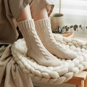 Può includere: Un paio di calzini color crema a trecce. I calzini spessi hanno un polsino a coste e un motivo a treccia. Sono appoggiati su una coperta bianca a maglia grossa.