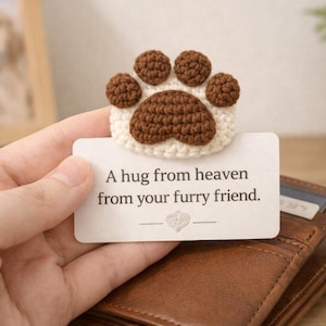 Pode incluir: Marcador de pata de crochê em marrom e creme, com o texto "A hug from heaven from your furry friend". O marcador é segurado sobre uma carteira de couro marrom. Foto emoldurada de um cachorro ao fundo.