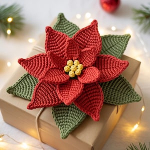 May include: A handmade crochet poinsettia flower in red, green, and yellow, resting on a brown gift box. The flower has detailed petals and leaves, with a yellow center. The box is tied with twine, suggesting a holiday theme.