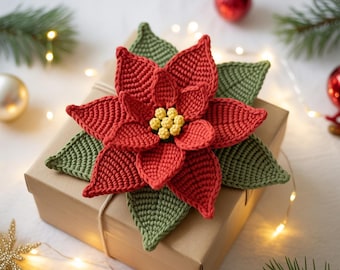 Pode incluir: Uma flor de poinsettia de crochê feita à mão em vermelho, verde e amarelo, repousando sobre uma caixa de presente marrom. A flor tem pétalas e folhas detalhadas, com um centro amarelo. A caixa é amarrada com barbante, sugerindo um tema de feriado.