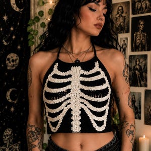 May include: A black halter-neck crop top featuring a white crocheted rib cage design on the front. The garment is constructed with a textured knit pattern, showcasing a central sternum and horizontal rib bones that extend across the chest area.