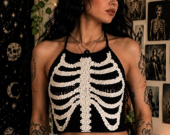 May include: A black halter-neck crop top featuring a white crocheted rib cage design on the front. The garment is constructed with a textured knit pattern, showcasing a central sternum and horizontal rib bones that extend across the chest area.