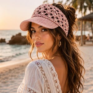Puede incluir: Una visera de ganchillo en color rosa claro con un ala estructurada y un patrón de tejido abierto adornado con pequeñas cuentas blancas. La visera está confeccionada con un material tejido e incluye una visera frontal sólida y curva.