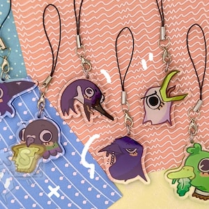 Pu&ograve; includere: Una collezione di ciondoli in acrilico a tema uccelli dei cartoni animati con cordini neri. I ciondoli presentano vari disegni di uccelli in viola, verde e bianco, alcuni con cibo. Lo sfondo ha un motivo rosa e blu.