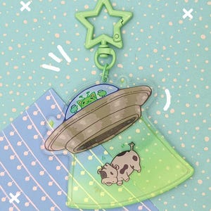 Może przedstawiać: Zielony brelok w kształcie gwiazdy przymocowany do przezroczystej akrylowej zawieszki. Zawieszka przedstawia kreskówkową ilustrację UFO z kosmitami i porwaną krową. UFO jest szare, a krowa brązowo-biała.
