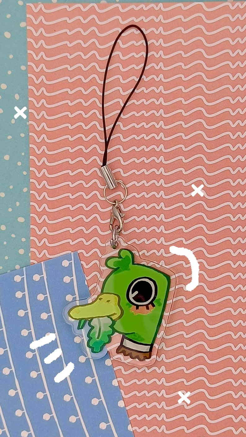 Pu&ograve; includere: Un ciondolo in acrilico a forma di anatra verde con cinturino marrone e chiusura argentata. Il ciondolo ha un design a cartone animato con un occhio grande e un becco giallo. Lo sfondo presenta un motivo a onde rosa e un motivo a pois blu.