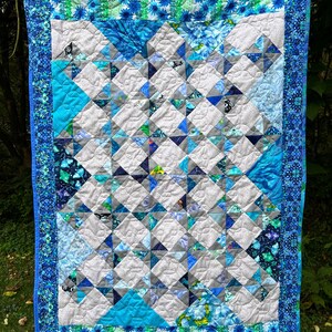 Op de afbeelding: Handgemaakte quilt in blauw- en grijstinten. Het heeft een geometrisch patroon van vierkanten en driehoeken, met een rand van bloemenstof. De quilt hangt aan een waslijn, wat de patchwork details benadrukt.