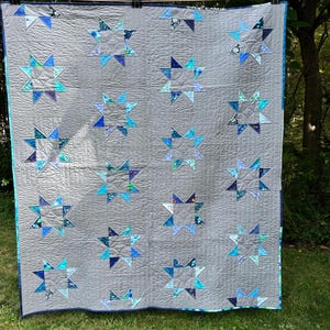 May include: A handmade quilt with a gray background and a repeating pattern of blue and teal star shapes. The stars are made of various patterned fabrics. The quilt is hanging outside on a clothesline.