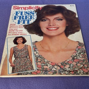 May include: A vintage Simplicity sewing pattern magazine cover featuring a woman with brown hair and a floral print dress. The magazine title reads "Fuss Free Fit". The cover includes a smaller image of a woman in a floral dress.