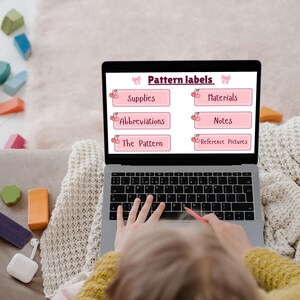 Op de afbeelding: Een laptop met "Pattern labels" met categorieën zoals "Supplies" en "Materials". Het scherm staat op de schoot van een persoon, met kleurrijke houten blokken en een deken in de buurt. De persoon gebruikt een roze potlood.
