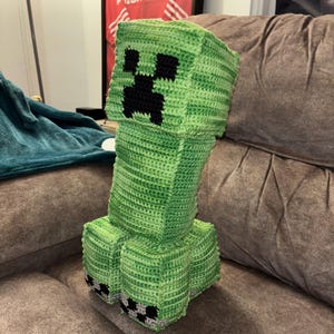 Pode incluir: Um Creeper verde de crochê feito à mão, inspirado no videogame Minecraft. A figura de pelúcia tem uma cabeça quadrada com um rosto preto pixelizado, um corpo longo e pés quadrados com detalhes em preto e cinza. Está em um sofá marrom.