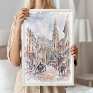 May include: A framed watercolour painting depicting a European city street scene with a tall gothic cathedral, multi-storey buildings, and a horse-drawn carriage. The artwork features soft blue, brown, and grey tones with pedestrians walking along the cobblestone path.