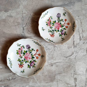 May include: Two white, scalloped-edge plates with floral designs. Each plate features a central arrangement of pink roses, purple buds, and green leaves, accented with orange berries and red details. The rims are trimmed with gold.