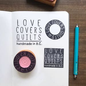 May include: Four black and white rubber stamps with the text "LOVE COVERS QUILTS handmade in B.C."  The stamps are on a white notebook page. One stamp is a circle with a heart inside. The other three stamps are rectangular.