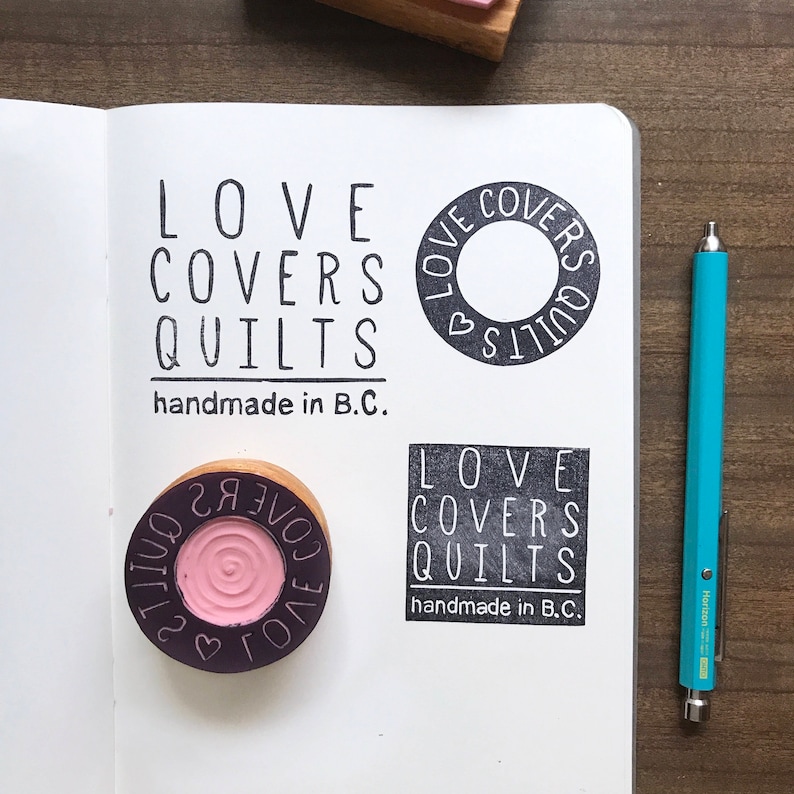 May include: Four black and white rubber stamps with the text "LOVE COVERS QUILTS handmade in B.C."  The stamps are on a white notebook page. One stamp is a circle with a heart inside. The other three stamps are rectangular.