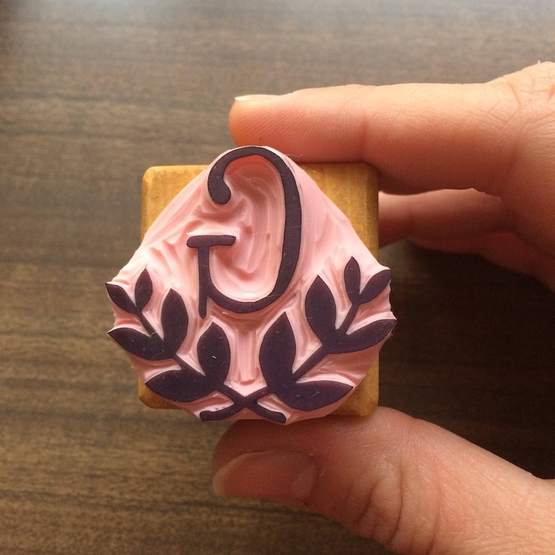 May include: A wooden block stamp with a pink rubber surface. The stamp features a black letter "G" surrounded by a wreath of leaves.