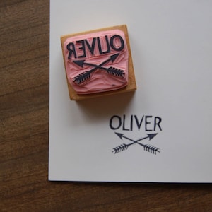May include: A wooden stamp with the name "OLIVER" in a bold, black font, with two crossed arrows beneath it. The stamp is pressed onto white paper, leaving a matching impression. The stamp's rubber surface is pink.
