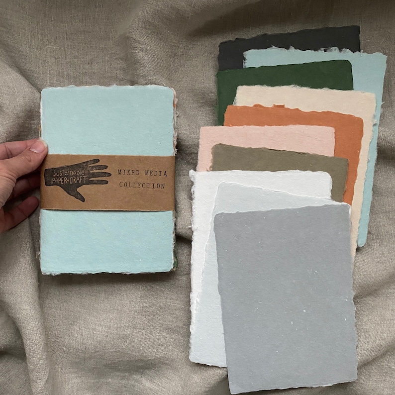 May include: A stack of handmade, textured paper sheets in various colors, including light blue, green, brown, and gray. The paper is labeled "Mixed Media Collection" and "Sustainable Papercraft".