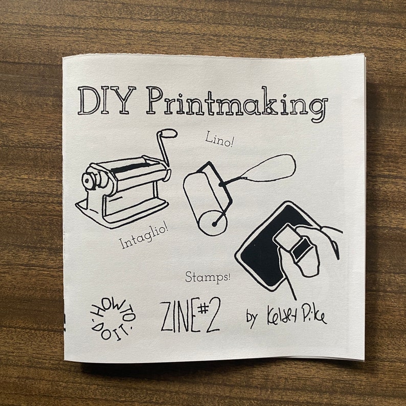 May include: A black and white illustration of a zine cover with the title "DIY Printmaking" and the text "ZINE #2 by Kelsey Pike". The cover features illustrations of printmaking tools, including a brayer, a printing press, and a stamp.
