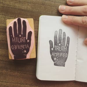 May include: A black and white handprint rubber stamp with the text "TRUTH AMPLIFIED" and small stars. The stamp is on a white page in a notebook.