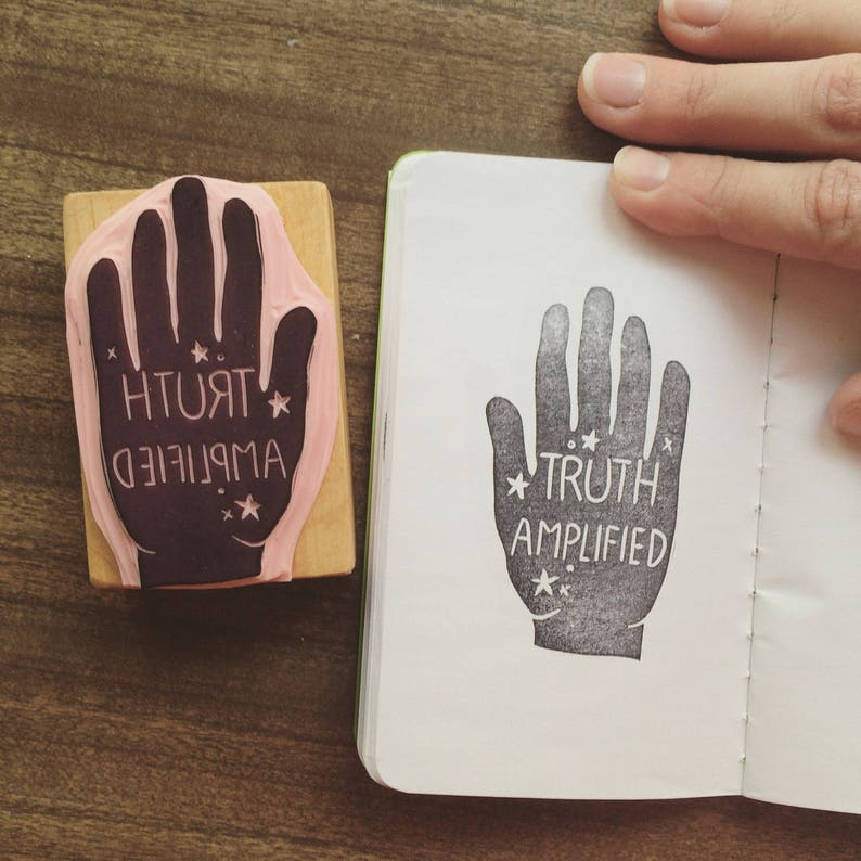 May include: A black and white handprint rubber stamp with the text "TRUTH AMPLIFIED" and small stars. The stamp is on a white page in a notebook.