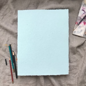May include: A light blue sheet of paper with a slightly rough texture, lying flat on a gray fabric surface. There are several pencils and a charcoal stick lying next to the paper.
