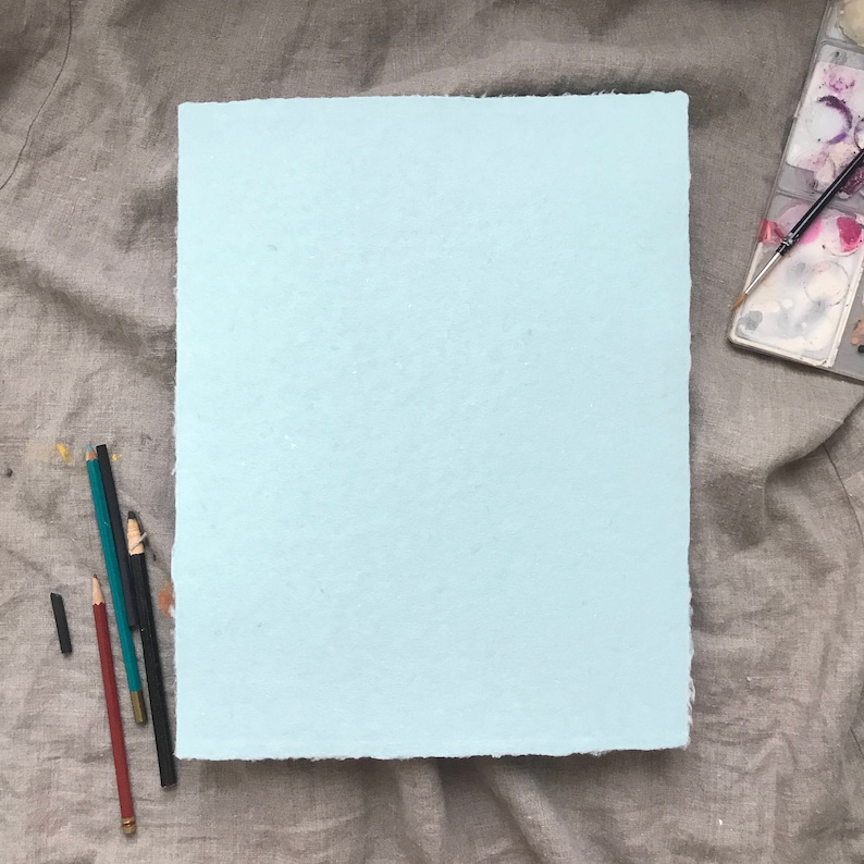 May include: A light blue sheet of paper with a slightly rough texture, lying flat on a gray fabric surface. There are several pencils and a charcoal stick lying next to the paper.