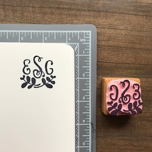 Puede incluir: Un sello de madera con fondo rosa y tinta negra. El sello presenta las letras "ESG" en una fuente cursiva con un diseño floral.