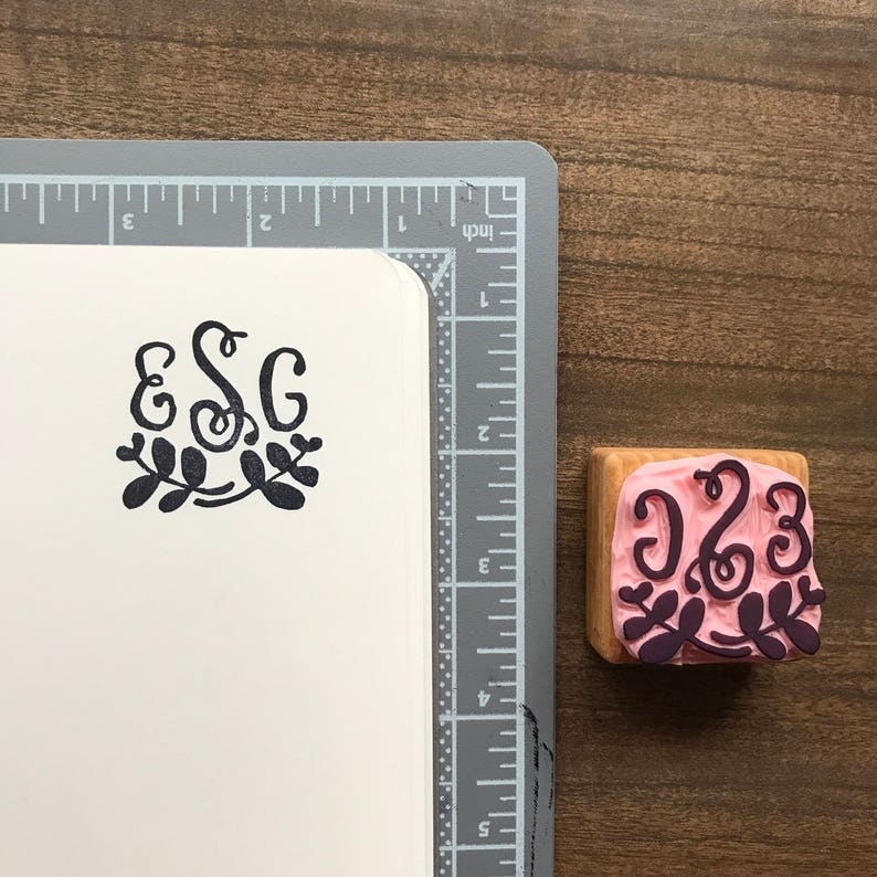 May include: A wooden stamp with a pink background and black ink. The stamp features the letters "ESG" in a cursive font with a floral design.