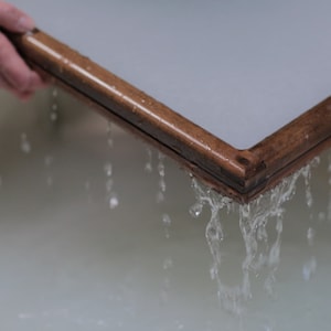 May include: A wooden frame with a screen is being lifted out of a basin of water. Water is dripping from the frame.
