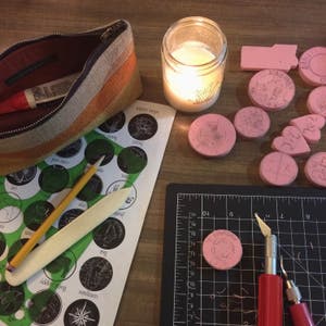 May include: A close-up of a craft table with a variety of tools and supplies for creating rubber stamps. The table includes a cutting mat, a craft knife, a ruler, a pencil, a wooden tool, and a variety of rubber stamps. The stamps are in various shapes and sizes, and some have designs on them. The table is covered in a brown wood surface.