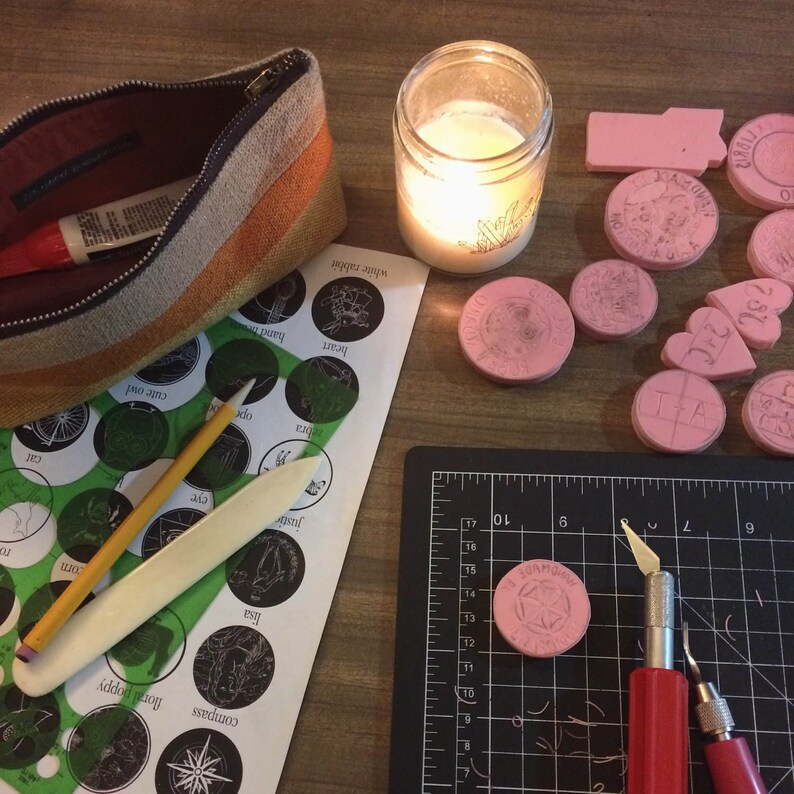 May include: A close-up of a craft table with a variety of tools and supplies for creating rubber stamps. The table includes a cutting mat, a craft knife, a ruler, a pencil, a wooden tool, and a variety of rubber stamps. The stamps are in various shapes and sizes, and some have designs on them. The table is covered in a brown wood surface.