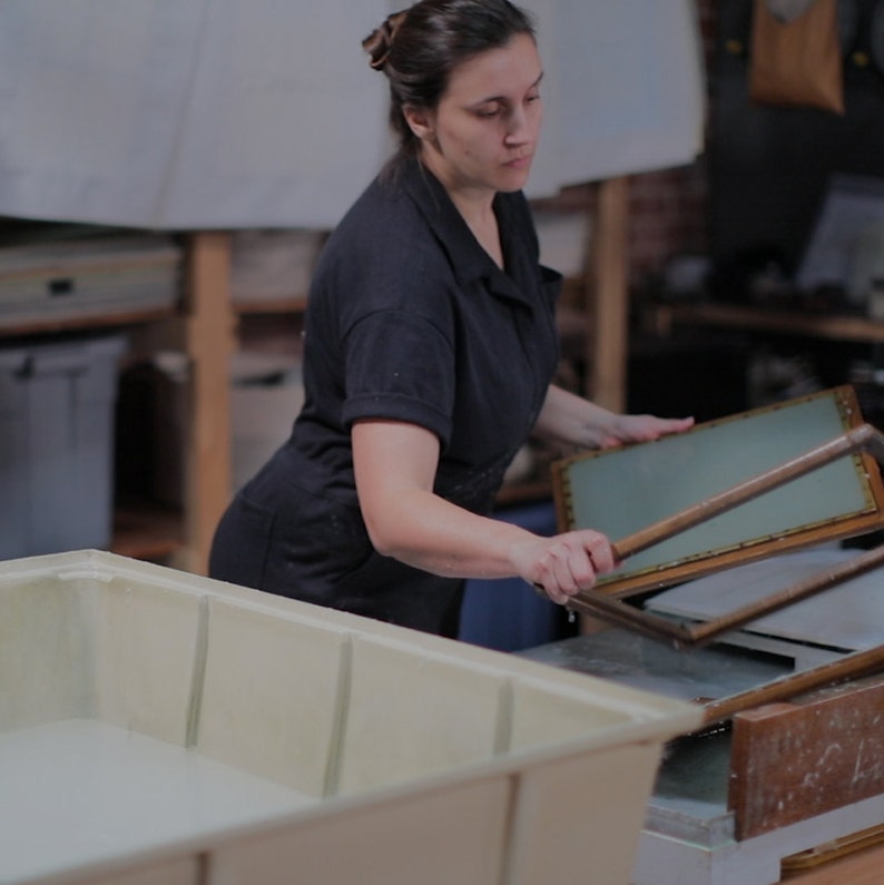 May include: A person using a wooden frame and a long wooden stick to press paper pulp in a large white tub. The frame is made of wood and has a metal mesh screen. The person is wearing a black shirt and is standing in a workshop.