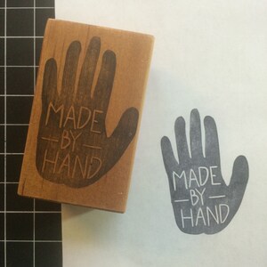 May include: A wooden hand stamp with the text "MADE BY HAND" printed on it. The stamp is used to create a gray handprint with the same text on a white surface.