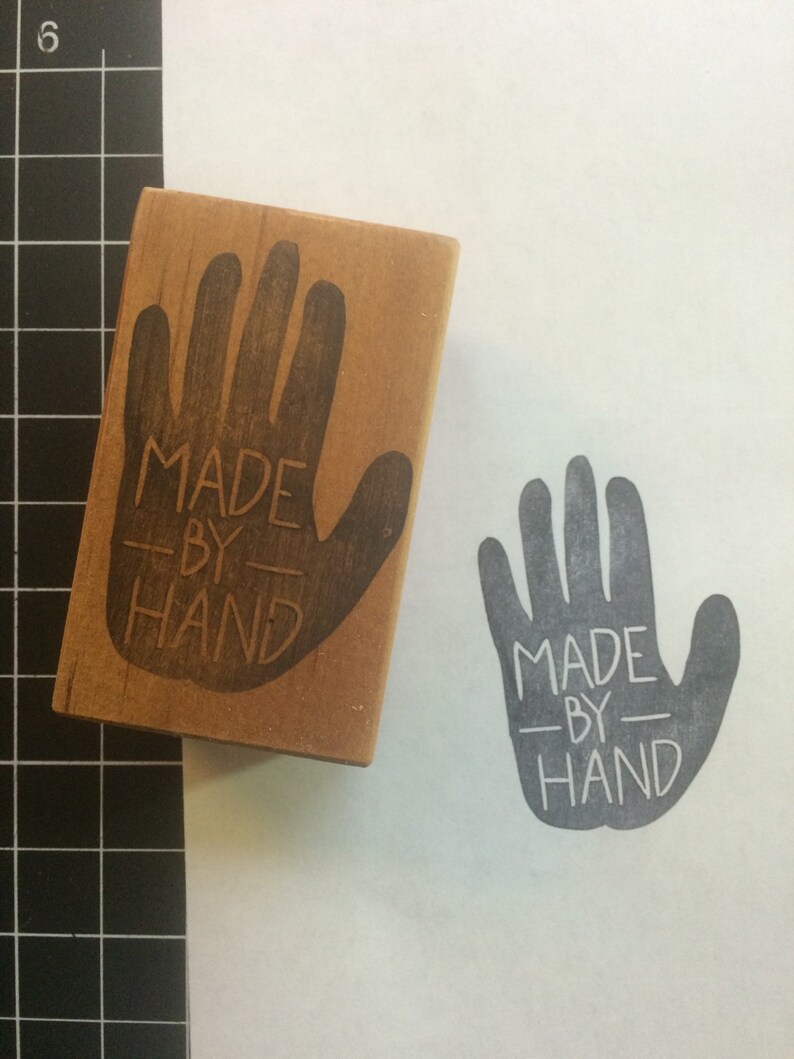 May include: A wooden hand stamp with the text "MADE BY HAND" printed on it. The stamp is used to create a gray handprint with the same text on a white surface.