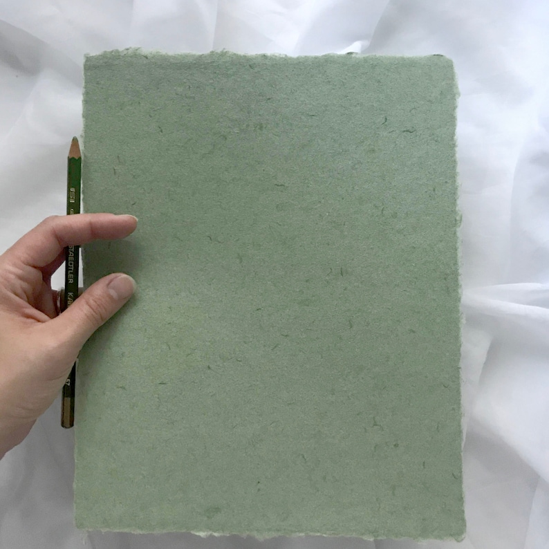 May include: A sheet of light green handmade paper with a rough, textured surface. The paper has a slightly irregular shape and is being held by a hand.