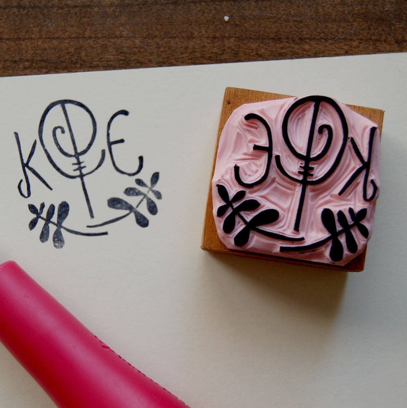 May include: A close-up of a pink rubber stamp with black ink. The stamp features a stylized design of the letters "KPE" with a floral motif. The stamp is on a white surface.