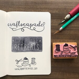 May include: A close-up of a wooden stamp with a pink rubber surface. The stamp features a design of a spool of thread, a can with the letters "XXX" on it, and a pair of scissors. The text "www.craftscapade.com" is printed on the bottom of the stamp.