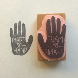 May include: A wooden stamp with a handprint design and the text "MADE BY HAND" in black lettering. The stamp is mounted on a wooden block.
