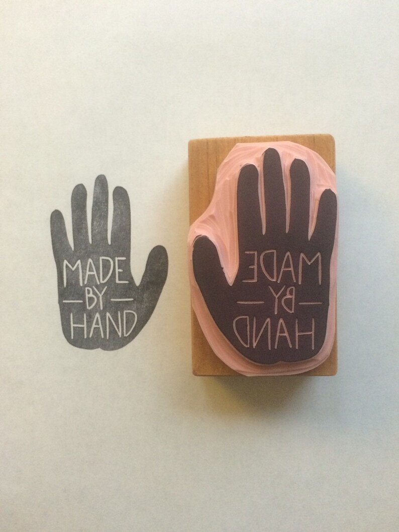 May include: A wooden stamp with a handprint design and the text "MADE BY HAND" in black lettering. The stamp is mounted on a wooden block.